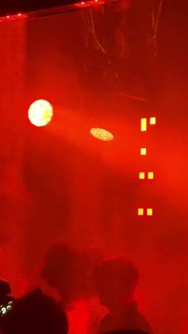 The video depicts a night club environment bathed in red light. The scene is somewhat obscured by smoke or haze, but a small group of people can be discerned in the foreground, with some wearing hats. Two round lights and a set of vertically arranged rectangular lights contribute to the intense red illumination. In the background, a person with long hair is visible, illuminated by the red light. The sound indicates music is playing, consistent with a nightclub setting.