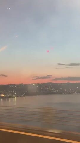 The video shows a view from a moving vehicle, likely a car, driving across a bridge at dusk or dawn. The sky is a gradient of colors, from a deep blue to a vibrant orange at the horizon. The body of water below reflects the city lights, creating a shimmering effect. The bridge railings are visible in the foreground, adding depth to the scene. The city skyline is dotted with numerous lights, outlining its silhouette against the colorful sky.