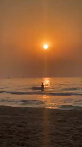 The video shows a beach at sunset, with a hazy orange sky above the ocean. The sun, a bright orange orb, is positioned high in the sky, casting a shimmering reflection on the water's surface. Small waves gently roll towards the sandy beach in the foreground. A silhouetted figure stands in the water, waist-deep, facing towards the sun. In the distance, two sailboats are visible on the horizon.