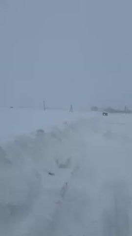 The video shows a snow-covered landscape. At the beginning of the video, we see a snowy path with tall snowbanks on either side.  In the distance, there are buildings.  The camera then pans up and shows a teal-colored tractor equipped with a snow plow.  The tractor is actively clearing a path through the heavy snow. A car is parked to the side of the tractor.