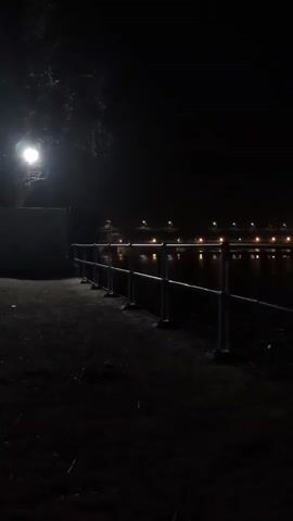 The video shows a dark scene at night next to a body of water. A lamppost illuminates a path and a metal railing that runs along the edge of the water. In the distance, lights from buildings or streetlights are visible across the water. The overall atmosphere is quiet and dimly lit.