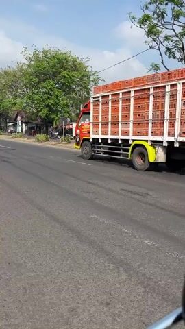 The video shows a road scene from the perspective of a vehicle following two trucks. The first truck, a bright yellow one, is in the foreground on the right side of the screen. It is moving forward on the road. The second truck, red and white, is in the background on the left side of the screen, also moving forward. A person on a dark-colored scooter, wearing a light purple jacket and a helmet, is in the right foreground, also moving forward on the road. The environment appears to be a sunny day with some trees and buildings visible along the side of the road in the background. The road is asphalt and has lane markings.