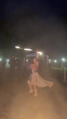 A woman fire dances at night in a low-lit outdoor area with a large tent and a crowd in the background. The woman wears a white dress with gold accents. She holds a double fire hoop, which she spins around her body. The fire from the hoop illuminates the area.