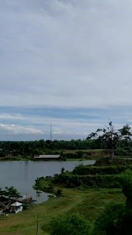The video shows a landscape view of a lake or pond surrounded by green vegetation and structures. The camera pans from right to left, revealing a dirt path on a hill overlooking the lake. In the distance, a town or city is visible under a partly cloudy sky. No people are visible in the video.