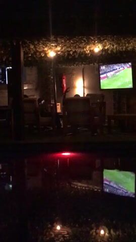 This nighttime video captures an outdoor or semi-outdoor patio area, likely a bar or restaurant, in what is contextually identified as Galadimawa, Nigeria. The scene is illuminated by string lights with round bulbs hanging from a leafy canopy overhead, casting a warm glow.

The main focus is a large flat-screen television mounted on a wall, displaying a football (soccer) match. Players in green and white jerseys are visible on the screen. Below this, a prominent reflection of the entire scene, including the TV and ambient lights, appears on a dark, reflective surface, possibly water or a very shiny floor. A distinct red light source is also reflected prominently in this lower surface.

In the background, several wooden tables and chairs are arranged, with dark silhouettes of people seated and observing the game. A red fire extinguisher is mounted on the wall to the left of the main television. The ambiance suggests a relaxed social setting for watching sports. No specific text or team names are clearly discernible on the TV screen, and the faces of the individuals present are not visible due to the low lighting and distance.