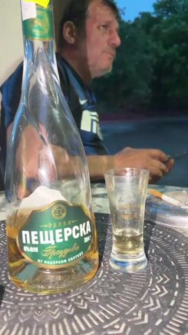 The image features a man in the background, out of focus, with a table in the foreground holding alcoholic beverages and smoking paraphernalia.

In the foreground, a distinctively shaped glass bottle of "Пещерска" (Peshtarska) rakia is prominently displayed. The label clearly states "РАКИЯ" (Rakia), "ПЕЩЕРСКА" (Peshtarska), and "ГРОЗДОВА" (Grozdova), indicating it is grape rakia. It is specified as "40% vol" and "700ml," produced "ОТ ПОДБРАНИ СОРТОВЕ" (from selected varieties). A yellow price sticker with "7.49 €" is attached to the bottle's neck.

Next to the bottle is a small shot glass, partially filled with a golden-brown liquid, and also bearing the "Пещерска" brand. An ashtray containing a lit cigarette rests beside the shot glass. The table surface features an intricate, textured pattern.

In the background, a man, likely in Sirakovo, Bulgaria, is seen from the chest up. He wears a dark blue and black striped short-sleeved shirt, resembling a sports jersey, possibly with a sponsor name like "IRELLI" (likely Pirelli). His head is turned to the right, and his mouth is slightly open, suggesting he is engaged in conversation. One hand is visible, holding what appears to be a mobile phone. The lighting suggests late afternoon or early evening. Green foliage is visible in the distant background, indicating an outdoor or semi-outdoor setting.