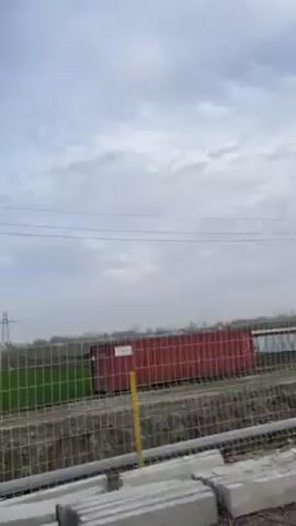 The video shows a drive-by view of a rural landscape, visible through a car window. A green field stretches to the horizon under a sky with thin, patchy clouds. Power lines run across the top of the frame. A utility tower stands in the distance. Along the side of the field, there’s a metal fence and some construction materials, including pipes and concrete blocks. A red shipping container is visible on the edge of the field. The camera is inside a vehicle, and it pans slightly, following the roadside. The scene appears to be a rural or semi-rural area undergoing some kind of construction or development.
