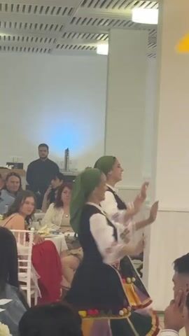 The video shows two young women in traditional folk costumes dancing in a brightly lit indoor space with tables and seated guests. The women are wearing green headscarves, white blouses with patterned sleeves, black vests, and colorful skirts adorned with tassels. They move in unison, their arms held out and hands making graceful gestures. Seated at tables around them are numerous people, some observing the dancers, others talking. The room has white walls and pillars, with a patterned ceiling and artificial lighting. A man in a black shirt stands near a serving station in the background. The camera moves slightly, following the dancers as they perform.