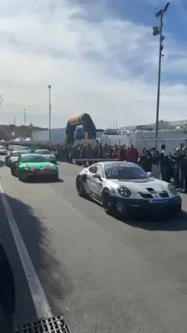 The video shows race cars slowly parading down a street. The first race car shown is teal, orange, and green with the number 52 on the side. The second car is silver with dark racing tires and the number 20 on the side. Many people are standing on the sidewalks on either side of the road, watching the cars. A large inflatable archway and other structures are visible in the background. The sky is blue with some clouds.