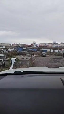 The video shows a view from inside a vehicle, looking out at a somewhat dilapidated urban landscape under an overcast sky. In the foreground, the dashboard and part of the windshield are visible, with streaks of water suggesting it might be raining or recently rained. A white side mirror extends from the left side of the vehicle.

Beyond the vehicle, the scene unfolds into a dirt and gravel area with some concrete structures. A silver sedan is parked in the middle ground. In the background, there is a collection of buildings of varying heights and colors, including what appear to be apartment buildings and smaller structures. Two prominent white, dome-shaped radar or weather station facilities are visible on a rise behind the buildings. The overall impression is one of a functional but perhaps remote or economically challenged settlement.