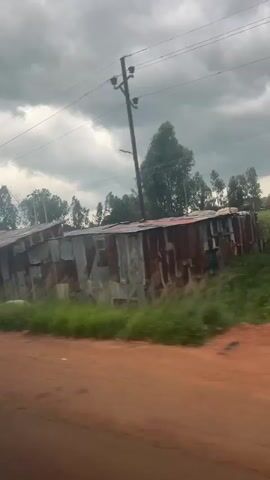 The video opens with a side-view shot of a rustic dwelling constructed with corrugated metal and wood. The structure appears weathered, with vegetation growing around its base and debris scattered nearby. The sky is overcast with heavy clouds. As the video progresses, the view shifts to a roadside scene. Large, white, opaque plastic bags, presumably filled with materials, are piled up along the side of a dirt road. Beyond the bags, a body of muddy, reddish-brown water is visible, crossed by a simple bridge with concrete supports and a metal railing. Trees and lush green grass line the banks of the water. The camera continues to pan, revealing more of the landscape, including more trees and what appear to be utility poles and wires overhead. The overall environment suggests a rural or semi-rural setting with some evidence of infrastructure and natural elements.
