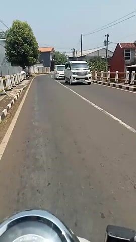 The video shows a person riding a motorcycle on a paved road. There are white barriers and a sidewalk on the left side of the road, with trees and buildings beyond. On the right side of the road, there are more buildings and vegetation. Several white vans and a black car are driving on the road. The weather appears to be sunny. The video is recorded from the perspective of the motorcycle rider.