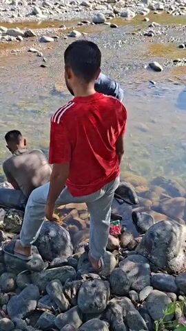 This media file captures a group of people by a river in Mane, Indonesia. The scene appears to be set in the daytime, with the sun casting shadows and illuminating the water and rocky riverbed. The weather seems pleasant, suitable for outdoor activities.

The primary subjects are several men, dressed casually in t-shirts and trousers or shorts. One man in a red t-shirt with white stripes is prominently featured, standing on the rocks near the water's edge. Another man, in a grey t-shirt, is seated on a large rock in the river. A third man, wearing a black t-shirt and a black cap, is seen walking near the water. A fourth man, wearing an orange t-shirt, is also visible. The group seems to be engaged in some activity near the river, possibly fishing or collecting something from the water, as indicated by the presence of a light blue bucket partially submerged.

The setting is a natural environment characterized by a clear river flowing over a bed of smooth, varied-sized rocks. The riverbanks are also rocky, with some sparse vegetation visible. In the background, a broader view of the river and the surrounding terrain can be seen, with more rocks and distant hills or mountains covered in lush greenery. The water is clear enough to see the rocks beneath the surface.

There is no discernible text within the media file. The overall mood seems relaxed and casual, suggesting a day out in nature.