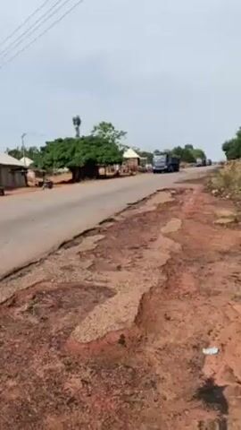 The video shows a rural scene with a damaged asphalt road. Several blue dump trucks drive by, followed by others in the distance. There are scattered buildings, trees, and a motorcycle in the background. The environment seems dry with brownish soil along the road edges. The sky is overcast.