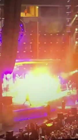 This video captures a live concert performance. The stage is illuminated with vibrant purple and blue lights, and pyrotechnics are active, creating a large burst of flame and smoke in the center of the stage. Several band members are visible on stage, playing instruments. In the foreground, a crowd of concertgoers is gathered, many holding up their phones to record the event. Above the stage, a raised platform is visible, with one person standing on it. The overall atmosphere appears energetic and dynamic.