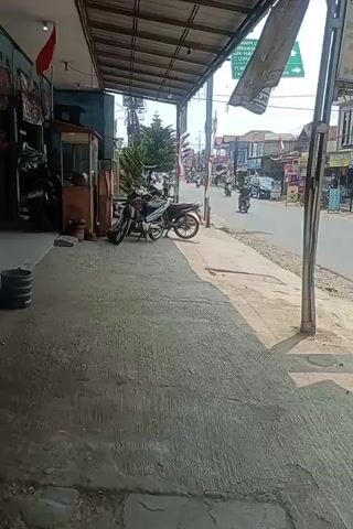 A street view shows a sidewalk next to a road. There are several motorcycles parked on the sidewalk, and some people are walking by. The road has cars and motorcycles driving on it. The buildings lining the street appear to be shops or businesses. There are some trees and utility poles visible. The sky is cloudy. The video appears to have been taken from a stationary position.