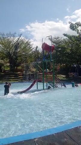 The video shows a water park with a blue pool and a red and yellow water slide. Several people are in the water, playing and sliding. The sun is shining, and the sky is blue with some clouds. Trees and greenery surround the pool.