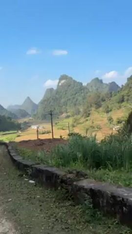 The video shows a landscape view from a low vantage point, possibly from a moving vehicle on a road. The foreground includes a dark concrete curb or barrier along the roadside, with some weeds growing near it. Beyond the curb, there's a field of brownish dirt or dry grass. The middle ground features a rural landscape with rice paddies or fields, some of which appear to be harvested, as well as small houses or structures. A power line runs across the scene. The background is dominated by a series of green, rugged mountains under a bright blue sky with some light clouds. The camera appears to be moving, as the view shifts slightly.