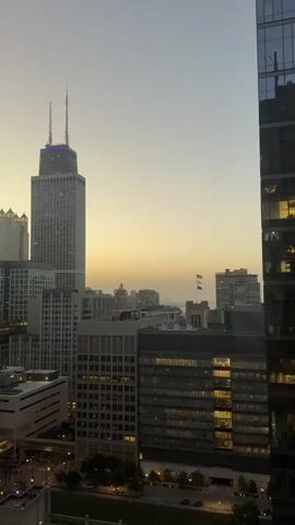 A slow zoom out reveals a cityscape at dusk. In the foreground, a tall, modern skyscraper with a glass facade is on the right. In the middle ground, a cluster of buildings, including a prominent, taller skyscraper with two antennae at the top, is visible. The sky above is a pale yellow and grey, indicating twilight. Lights are visible in some of the windows of the buildings. The overall impression is of a serene urban environment as day transitions into night.