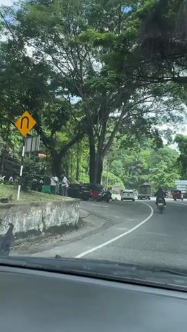 The video shows a view from inside a car driving on a road in a lush, green area. The road curves slightly to the right. A white car is parked on the left side of the road. A person on a motorcycle is driving along the road. In the distance, other vehicles, including a truck and a tuk-tuk, can be seen. Several people are standing near the parked cars on the left side of the road. The area is surrounded by trees and greenery, suggesting a natural or rural setting. The sky is partially cloudy, and the overall lighting is bright.