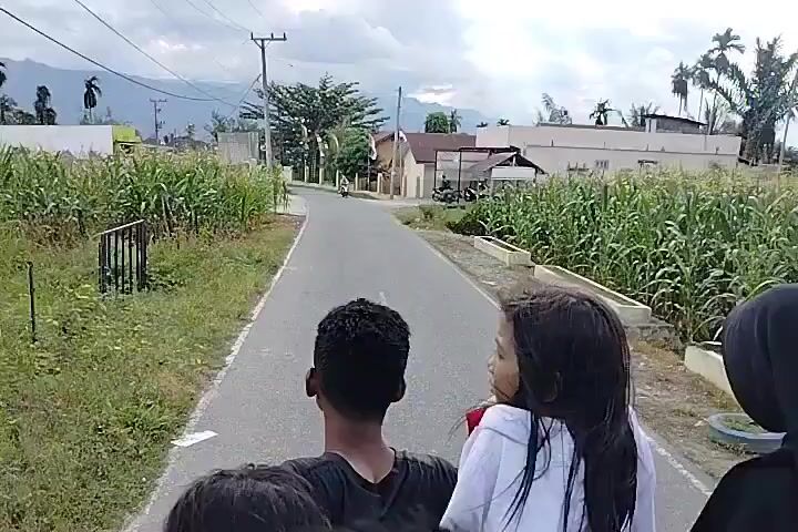 A point-of-view shot from the back of a moving vehicle shows three people, two adults and one child, riding on the back. The driver of the vehicle is not visible. The child, a girl with long dark hair, is looking to the right, while the two adults are facing forward. The vehicle is driving down a paved road in a rural area. On either side of the road are fields of corn. In the distance, there are houses and mountains. A motorcycle is also visible on the road ahead.