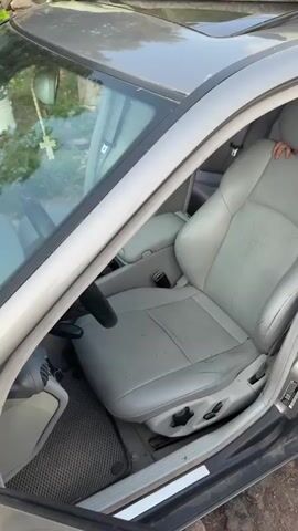 The video shows the exterior of a silver Mercedes-Benz car, with the driver's side door open. The interior of the car is visible, including the leather seats, steering wheel, and a rosary hanging from the rearview mirror. The car appears to be parked outdoors, with a building and some greenery in the background. The hood of the car has a sticker that says "Sportline" and "Produced by AMG".
