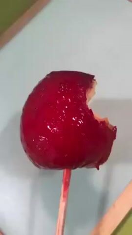 The video shows a close-up of a red candy apple on a wooden stick. A bite has been taken out of it, revealing the white apple inside. The apple sits on a light blue surface, which appears to be part of a framed object. A portion of another wooden stick is visible at the bottom right. The camera rotates to reveal the apple’s core. Then, the camera rotates back to its initial position.