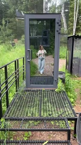 A young woman with shoulder-length blonde hair, wearing a light green crop top and a white cardigan with pink floral embellishments, along with light blue wide-leg jeans and white sneakers, stands on a metal grate platform in front of a mirrored glass door. She is holding a yellow smartphone and taking a selfie. The environment is outdoors with lush green grass and trees visible in the background. The mirrored door reflects the woman and the surrounding nature.