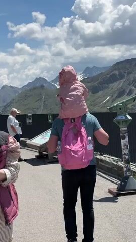 A person walks along a paved path carrying a child on their shoulders. The person is wearing a teal t-shirt, black pants, and a pink backpack with two water bottles in the side pockets. The child is wearing a pink hooded jacket with a white pattern. In the background, there are mountains, some with snow-capped peaks, under a partly cloudy sky. To the left, a man wearing a white hat and blue shorts is standing. To the right, a large green telescope is mounted on a pedestal with various stickers attached. The path is bordered by a dark metal fence.
