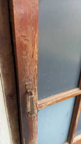 The video shows a person opening a rustic wooden door to reveal a small, utilitarian bathroom. The exterior of the door frame is made of weathered wood with a simple metal handle.

Upon opening, the interior of the bathroom comes into view. The walls are covered in dark, possibly black or dark blue, horizontal-ridged tiles. A small, rectangular window is positioned high on one wall, letting in natural light. The main fixture is a white squat toilet, set into a tiled floor with a mix of grey and beige square tiles. To the right of the toilet, a pink plastic jug and a yellow plastic bucket are visible on the floor, suggesting water for flushing or washing. The scene depicts a simple, functional toilet, typical of those found in rural or semi-rural areas, such as Pirveli Kesalo, Georgia.