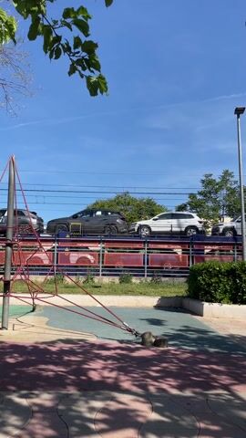 The video shows a train transporting cars passing by a playground on a sunny day. The train, loaded with various gray and white SUVs, moves from right to left in the frame. In the foreground is a children's playground with climbing equipment constructed of rope. The playground is situated on a surface with various color sections. Between the playground and the train tracks, there is a low hedge. Trees frame the scene on both sides, their leaves providing dappled shade on the ground. The sky is a clear, bright blue.