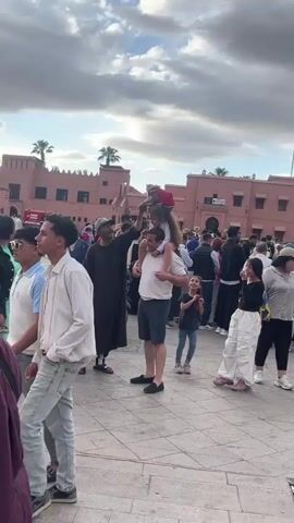 The video shows a public square, presumably in a Moroccan city. A man with a brown robe and a cap stands next to a man carrying a young girl on his shoulders. The man in the robe is holding a small monkey on a leash, lifting it up for the girl to see. Another young girl stands nearby, looking on with interest. People are walking around in the background. The buildings in the background are made of light-red brick, with palm trees visible in the distance. The sky is cloudy. The ground is paved with tiles. A red Moroccan flag can be seen on the rooftop of one of the buildings.