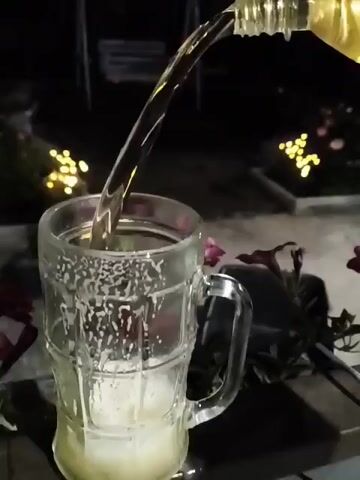 A stream of light-colored liquid is poured from an unseen container into a clear glass mug. The liquid fills the mug, creating a frothy head. The mug has a handle and vertical ridges. The background is dark with blurred yellow lights and some dark foliage.