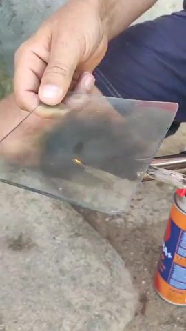 A person is holding a thin piece of glass in their left hand and a stick in their right hand. They are using a small blowtorch, attached to an orange can, to heat the end of the stick, which is placed against the edge of the glass. The flame from the blowtorch is visible and is directed at the stick. The person appears to be sitting down, with their legs crossed. They are wearing dark shorts and white clogs. The background is a light-colored, possibly concrete surface. The action suggests an attempt to melt or shape the glass with the heat from the stick.