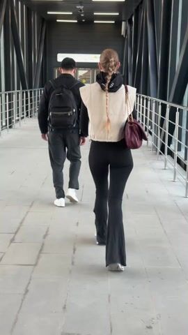 Two people walk along a pedestrian overpass. A woman with blonde braided hair walks ahead of a man. Both are viewed from behind. The man wears a black backpack, black clothing, and white shoes. The woman wears a black long-sleeved shirt, a cream-colored vest, black flared leggings, and a maroon purse. The overpass has a gray floor and silver railings. There are signs with red lettering. Through the windows of the overpass, angled black beams are visible.