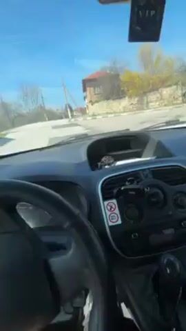 A first-person perspective video shows someone driving through a rural neighborhood on a sunny day. The dashboard and steering wheel of the vehicle are visible. The driver's left hand is mostly seen on the steering wheel. Outside the car window, there are houses with stone walls, trees with yellow foliage, utility poles, and another car further down the road. The sky is clear and blue.