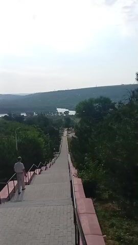 A person walks down a long, wide set of stairs. The stairs are paved with light-colored bricks and have a red stone railing. Trees and green foliage line the stairs on both sides. Beyond the trees, a body of water is visible, with rolling hills and a hazy sky in the background. The person is wearing light-colored pants and a light-colored shirt and appears to be walking away from the camera. The video is shot from a high angle, looking down the stairs. The overall impression is one of a scenic overlook or park.