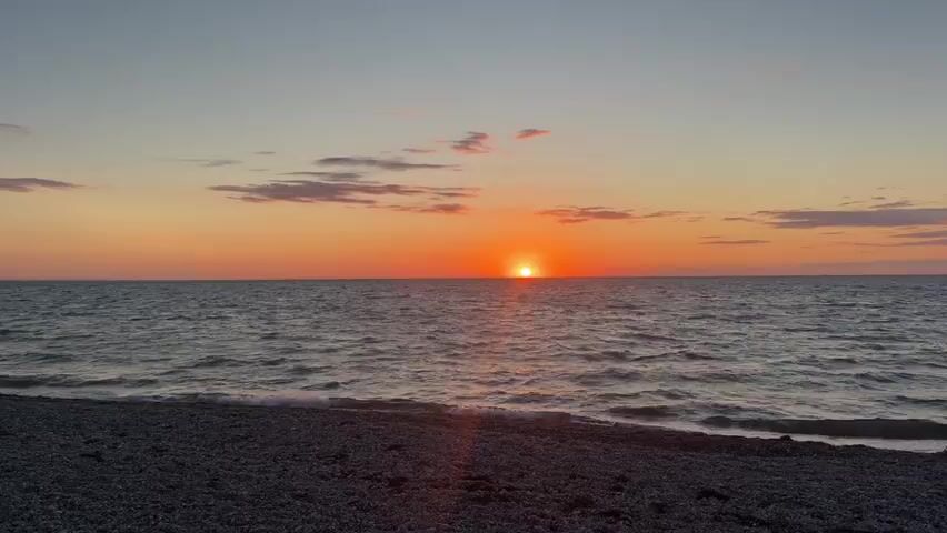 A wide shot captures a serene sunset over a calm ocean from a pebbled beach. The sun, a brilliant orange disk, hovers just above the horizon, casting a warm glow across the sky. Soft, elongated clouds streak the upper atmosphere in hues of pink and gray, contrasting with the deepening blue of the sky above them. The water, a dark, rippling expanse, reflects the sunset's colors, with small waves gently lapping against the shore. The beach itself is a textured carpet of various-sized gray and white pebbles, with the shoreline faintly visible in the foreground. There are no people or man-made objects immediately visible in the frame, emphasizing the natural beauty of the scene.