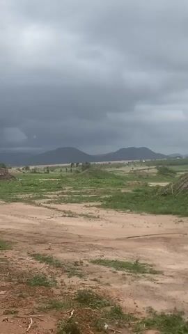 The video presents a panoramic view of a vast, open landscape under a heavily overcast sky. The camera slowly pans from left to right, revealing the expanse of the environment.

In the foreground and midground, there is mostly dry, reddish-brown dirt or soil, suggestive of a rural road or undeveloped land. Scattered patches of green grass and low-lying vegetation are visible across the ground, particularly in the middle distance. There are also some small mounds of earth or debris.

In the background, a series of dark, undulating hills or mountains stretch across the horizon, appearing hazy under the dense cloud cover. A few indistinct trees or structures can be faintly seen in the middle distance, blending into the landscape.

The sky is dominated by thick, dark grey clouds, indicating overcast weather, possibly before rain or during a heavily clouded day. The ambient sound includes the distinct whooshing of wind, which also appears to sway some of the distant foliage.

No people or specific activities are visible in the frame. The scene conveys a sense of untouched or sparsely developed natural surroundings. Given the provided context, this landscape is likely in Gosa, Nigeria, showcasing its natural terrain and weather conditions. There is no visible text.