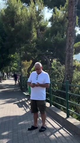 A man wearing a white t-shirt and khaki shorts stands on a paved walkway next to a metal railing. He is looking down at his phone and holding another object in his hand. The walkway is bordered by a row of trees and shrubs, and in the background, a person is walking away down the path. The sun is shining, casting shadows on the ground.