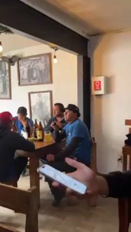 Here is a detailed description of the video.

The video shows the interior of a restaurant or bar. Several men are sitting around a wooden table, drinking beer. One man at the end of the table is looking at his cell phone. The table has an assortment of bottles of beer and limes, and the men are seated in wooden chairs. 

Behind the men are several framed pictures on the wall. There is a white first aid kit mounted on the wall as well.  A person in the foreground holds a smartphone up. 

The camera pans across the room, past another table, to the bar where a woman with curly hair stands. She appears to be looking directly at the camera. A television is mounted above the bar, and beer bottles are lined up behind her.

The bar area is decorated with greenery and hanging light bulbs. There is a window next to the bar area.