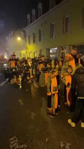 The video shows a street parade at night, with a group of musicians dressed in orange and black costumes playing drums on wheeled carts. The street is lined with spectators, including a child wearing an orange scarf and a woman carrying a silver platter of food. The buildings along the street are illuminated by streetlights, and there is some smoke or fog in the air. Debris is visible on the wet street. The overall atmosphere appears festive and lively.