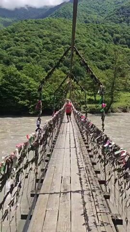 A person wearing a red tank top with "BULLS 91" and dark shorts walks or jogs across a wooden suspension bridge, moving towards the camera. The bridge, constructed with wooden planks and metal cables, is heavily decorated with numerous colorful ribbons and pieces of fabric tied along its railings and support wires. Beneath the bridge, a wide river with turbid, brownish water flows. In the background, large mountains covered in dense green trees rise under a partly cloudy sky. The setting is a natural, mountainous, and riverine landscape, characteristic of Bzypta, Abkhazia, during what appears to be a bright, sunny day. The person appears to be enjoying their walk on the swaying bridge.