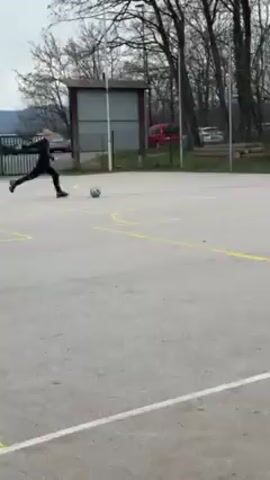 Two children practice soccer on a concrete court in an outdoor, wooded area. One child stands in the soccer goal while the other stands some distance away, lines up and kicks the ball toward the goal. The first child dives forward but the ball sails to the right. The first child stands and picks up the ball. The second child walks toward the goal.