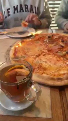 A close-up shot shows a partially eaten pizza with a variety of toppings including mushrooms, ham, artichoke hearts, and olives. Next to the pizza is a clear glass mug filled with dark liquid and slices of citrus fruit, likely tea. The mug sits on a small white saucer. Two people are visible in the background, sitting at the same wooden table. One person is using a knife and fork to cut a slice of pizza. The other person is partially obscured. The scene is indoors, likely a restaurant or cafe. The lighting appears natural.