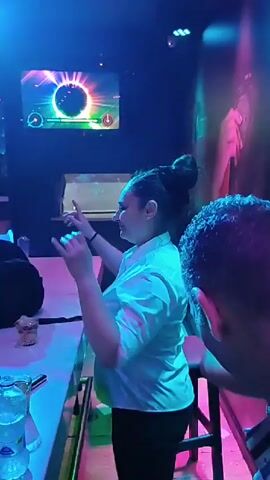 Here’s a detailed description of the video:

The video shows a brightly lit bar with colorful lights, focusing on bartenders and customers. Initially, a woman in a white top and dark pants is seen dancing behind the bar, raising her arms. A man sits at the bar to her left. 

The camera then pans across the bar, showing various bottles and glasses. More customers are visible at tables in the background. Several individuals are seated at the bar, and at one point, they appear to be applauding. Other bartenders, one in a black top and white shorts, briefly interacts behind the bar. Another woman, with red hair pulled up into a bun, is also seen working behind the bar. A television screen displays colorful graphics on the wall. The general environment suggests a lively, social setting, with people enjoying the bar's atmosphere and music.