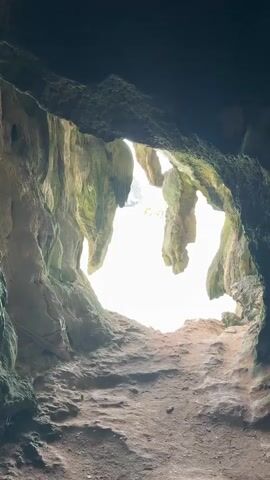 A vertical video opens from inside a cave, showing the rough, textured walls and ceiling of the cave. The view then pans to the right and slightly down, revealing a bright opening that leads to the outside. Through the opening, a body of water, appearing as a light green sea, is visible. In the distance, several islands are faintly seen, with some greenery on them. The cave's interior is dark, creating a stark contrast with the bright light from the exit. The ground inside the cave appears to be made of dirt and rocks. The camera continues to move, offering a wider view of the sea and islands. The video concludes by showing the exit of the cave framing the ocean view.