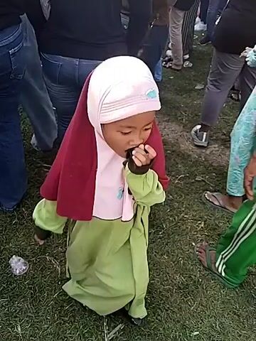 The video shows a gathering of people, seemingly at an outdoor event or celebration. A pink and green canopy provides shade for a seated audience. Several people are standing and walking around. There's a videographer with a tripod. The setting appears to be on a grassy field, with a rural or agricultural landscape visible in the background. Several people in the crowd are wearing headscarves.