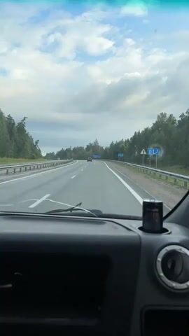 The video shows a first-person perspective from inside a vehicle, looking out the windshield and side window. The vehicle is traveling on a highway with multiple lanes in a wooded area. The sky is cloudy with patches of blue. Traffic signs are visible along the side of the road. A person's reflection can be seen in the side mirror, holding a phone, possibly taking a photo or video. The dashboard of the vehicle is visible in the foreground. The overall impression is of a journey on a highway.