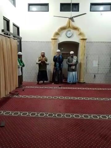 Four men are standing in a mosque with red carpet. The men are wearing traditional Muslim attire. The man in the center is speaking into a microphone. Another man is standing to his right, holding a book. A man on the far left is standing near a curtain. The man on the far right is standing with his arms crossed. The mosque has a clock on the wall.